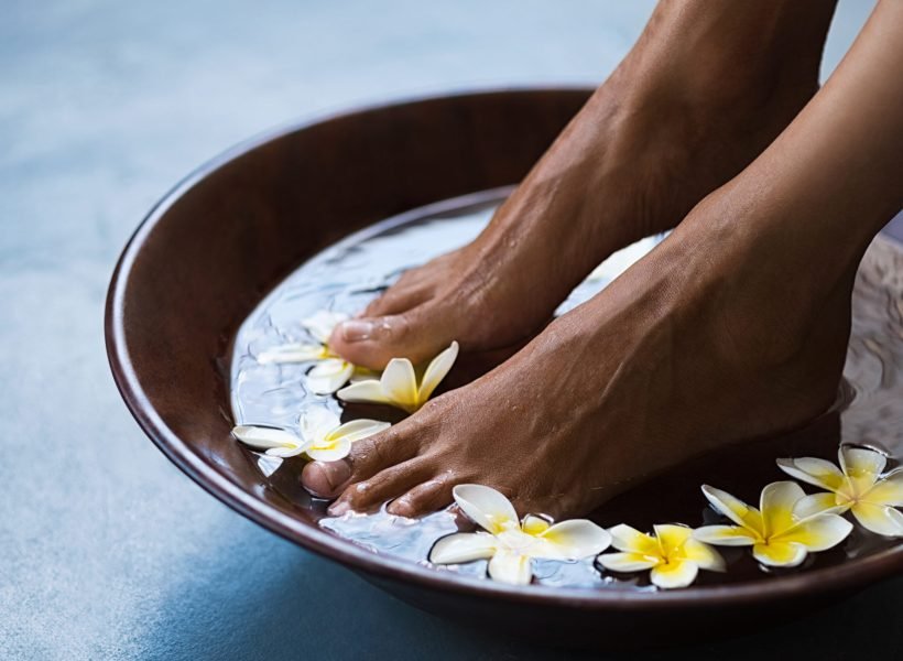 Woman soaking feet in bowl of water with floating frangipani flowers at spa. Closeup of a female feet at wellness center on pedicure procedure. Woman feet in spa wooden bowl with exotic white flowers.